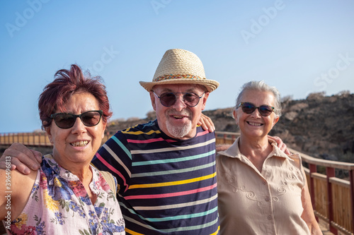 A group of three carefree elderly people looking at camera enjoying the seaside excursion standing in a wooden footpath. Active lifestyle for three retirees
