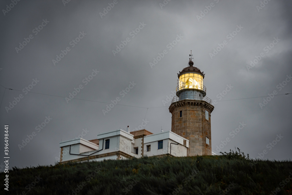 old lighthouse on a coastal hill of a small town, the lighthouse has a yellow flashing light, it is night and the sky is dramatic, gray with clouds