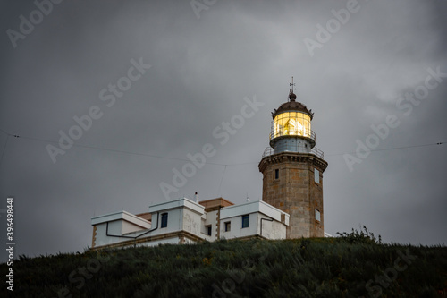 old lighthouse on a coastal hill of a small town, the lighthouse has a yellow flashing light, it is night and the sky is dramatic, gray with clouds