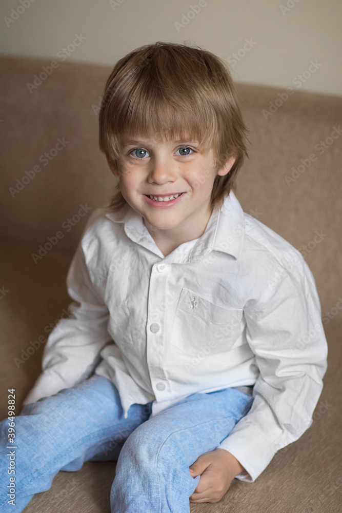 A five-year-old boy in a white shirt and blue pants is sitting in a beige room A five-year-old boy in a white shirt and blue pants is sitting in a beige room