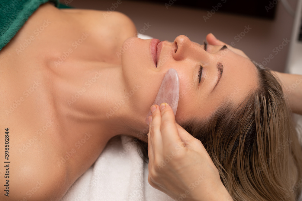 Young and beautiful woman during Chinese traditional massage - Gua Sha. Close-up photo. Beauty tools in the hand of beautican Beauty treatment in SPA salon