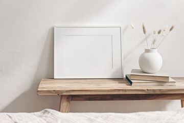 Winter still life. Horizontal white frame mockup on vintage wooden bench, table. Modern white ceramic vase with pine tree branches, Christmas paper ornaments and books. White wall background.
