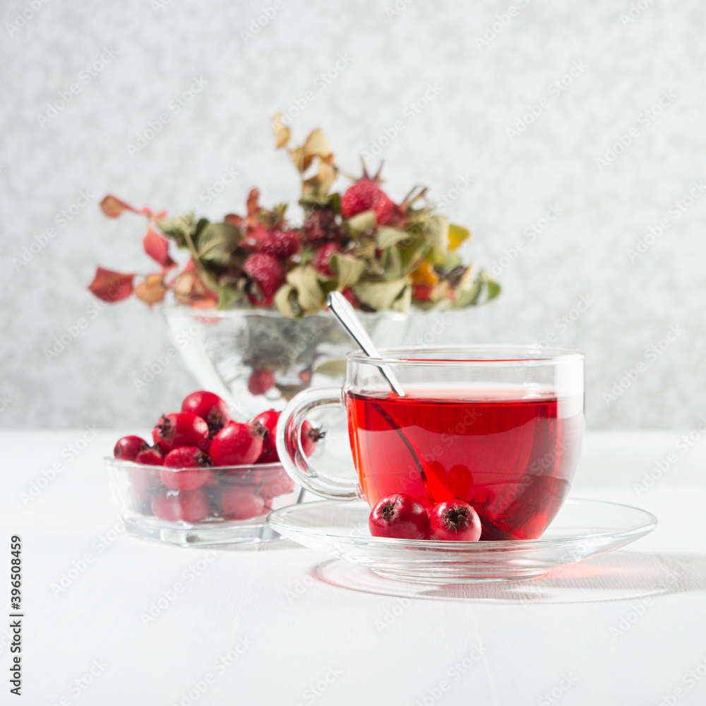 Autumn red herbal tea in transparent cup, spoon with hawthorn, rosehip berry, dried leaves in sun beam in soft light white interior, square. Autumn red herbal tea in transparent cup, spoon with hawthorn, rosehip berry, dried leaves in sun beam in soft light white interior, square.
