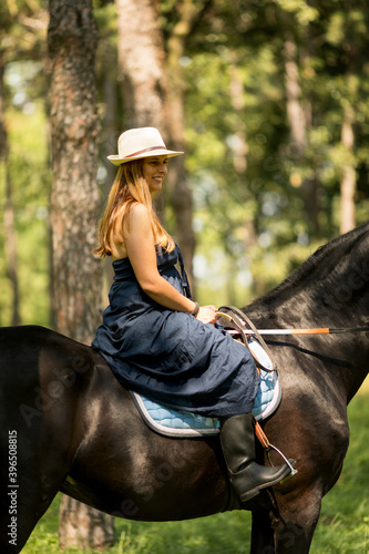 Girl with a long copper hair and long flowy shoulderless dress, ride a black horse with trees in the background.