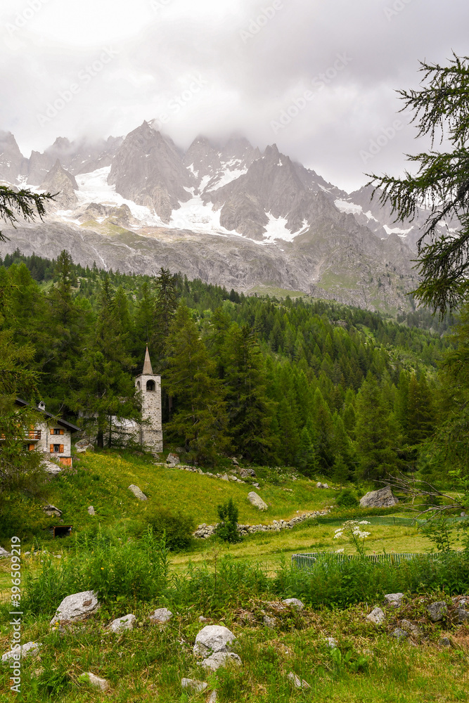A small stone church in a pine forest at the foot of the Mont Blanc massif in a cloudy summer day, Ferret Valley, Courmayeur, Aosta Valley, Alps, Italy
