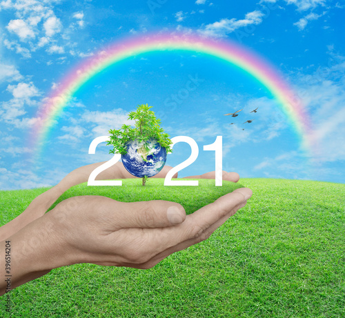 2021 white text with planet and tree on green grass field in man hands over blue sky, white clouds, and rainbow, Happy new year 2021 ecological cover, Save the earth concept, Elements of this image fu