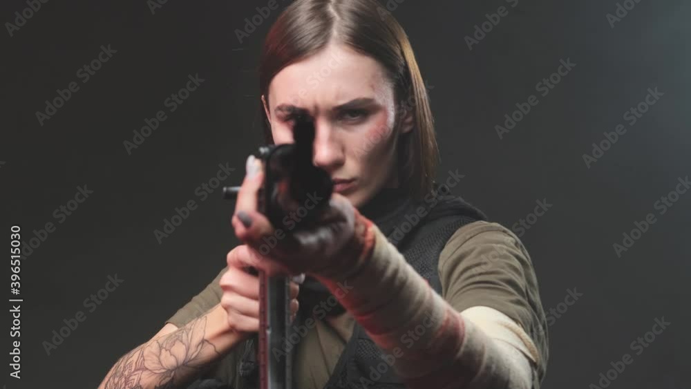 Dangerous and angry military woman in black vest with bandaged arm and short haircut aims a rifle looking at camera in dark background. Dangerous and angry military woman in black vest with bandaged arm and short haircut aims a rifle looking at camera in dark background.