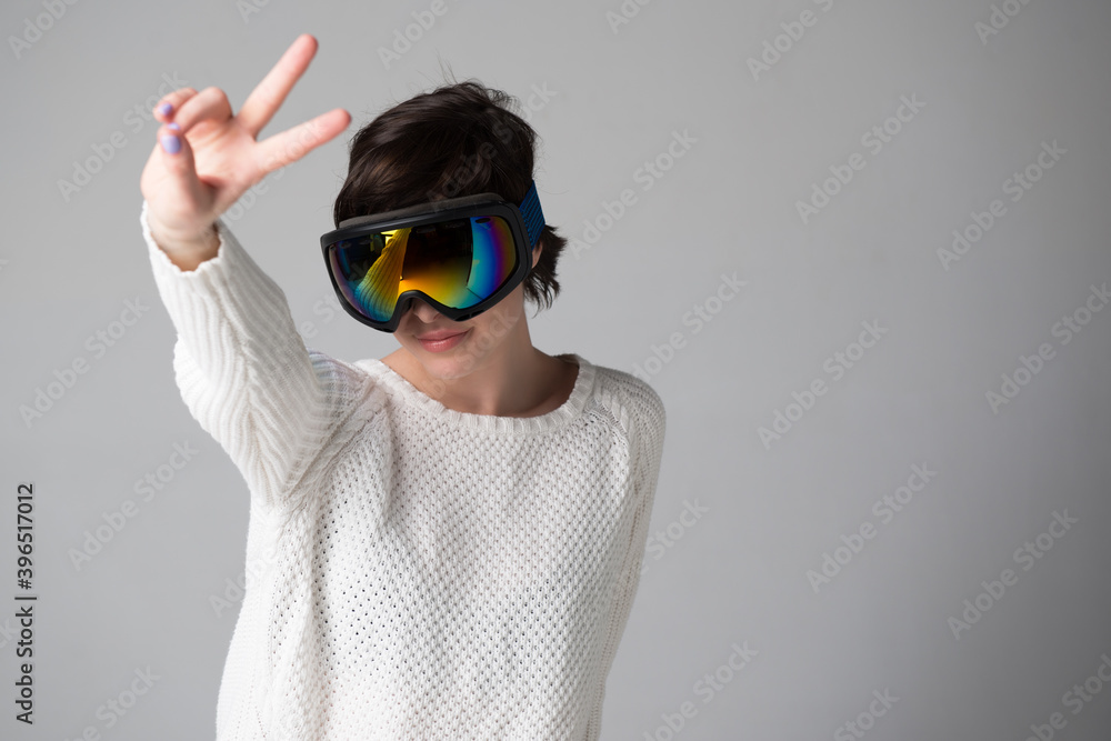 Female portrait in ski goggles with positive expressions and victory sign, winning gesture over gray background, snow winter sport equipment theme Female portrait in ski goggles with positive expressions and victory sign, winning gesture over gray background, snow winter sport equipment theme