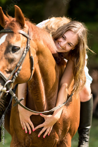 Girl with a long brown hair and tall boots rides a horse. She is leaning down and smiling, putting both hands around the horse lovingly, forming a heart shape with her fingers.