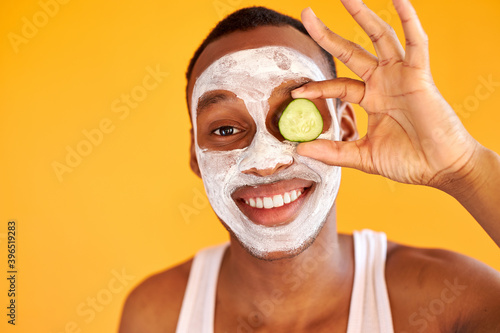 postive man apply face mask with cucumber slices, express positive emotions isolated over yellow background, male get beauty treatments