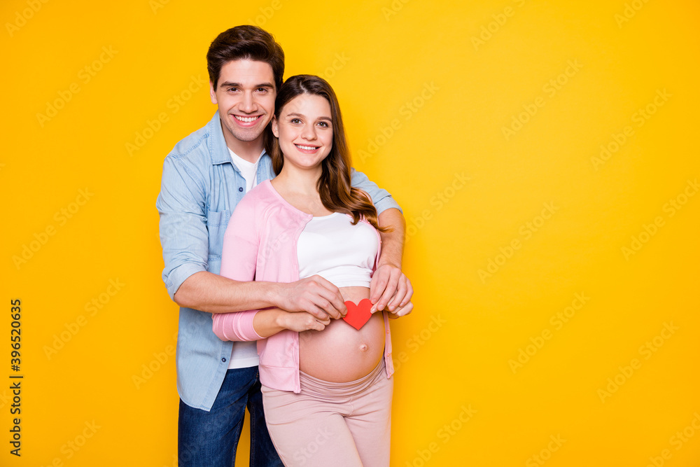 Photo of young couple stand hug pregnant woman show paper heart wear casual cloth isolated on yellow background Photo of young couple stand hug pregnant woman show paper heart wear casual cloth isolated on yellow background