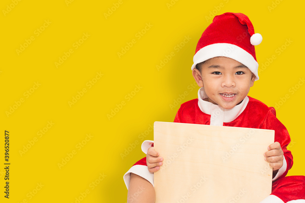Happy child wearing a santa claus costume with a wooden sign To enter text A picture of a boy with smiling against a yellow background. with clipping path. Happy child wearing a santa claus costume with a wooden sign To enter text A picture of a boy with smiling against a yellow background. with clipping path.