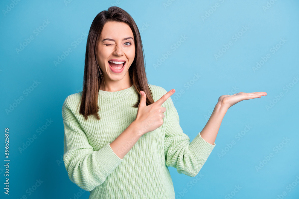 Photo of pretty young girl indicate finger arm hold empty space wink eye open mouth wear green sweater isolated blue color background Photo of pretty young girl indicate finger arm hold empty space wink eye open mouth wear green sweater isolated blue color background