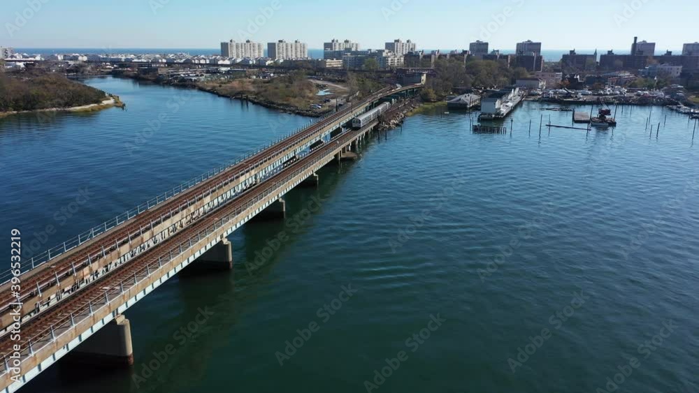 An aerial shot over Grassy Bay in Queens, NY. The camera dolly in behind a train on elevated tracks as it heads into the train yard. It is a beautiful day and the sun is shining on the water. An aerial shot over Grassy Bay in Queens, NY. The camera dolly in behind a train on elevated tracks as it heads into the train yard. It is a beautiful day and the sun is shining on the water.