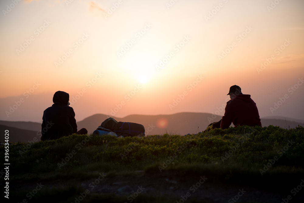 Two Hikers Sitting On The Mountain Ridge, Silhouette Of Resting Travelers Enjoying Sunset / Nature, People