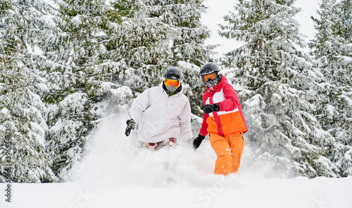 Man and woman in ski suits and helmets spending time together at ski resort, running through powder snow in winter forest with snowy coniferous trees on background. Concept of active leisure.