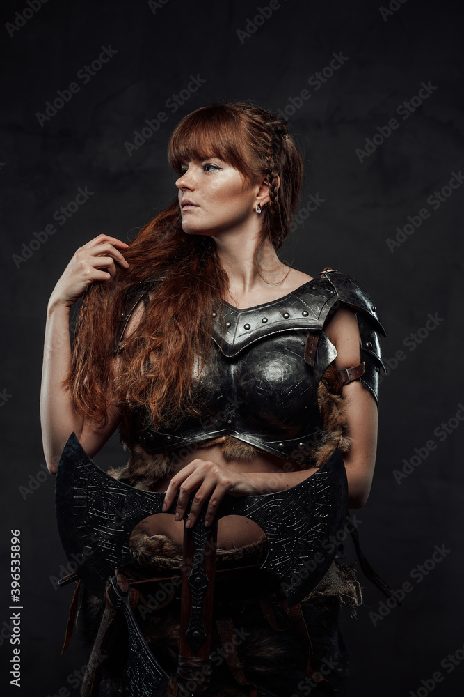 Naklejka premium Beautiful but dangerous warlike woman viking with long brown hairs in dark armour holding huge axe in dark background.