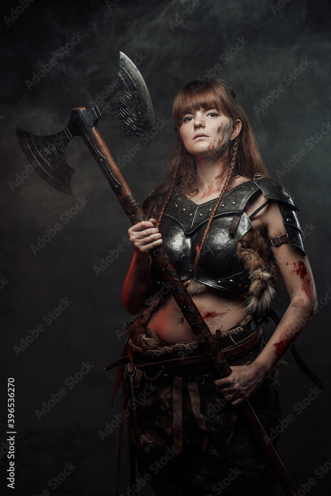 Armed with two handed axe nordic grimy amazon in dark armour with brown hairs poses in dark background with smoke. Armed with two handed axe nordic grimy amazon in dark armour with brown hairs poses in dark background with smoke.
