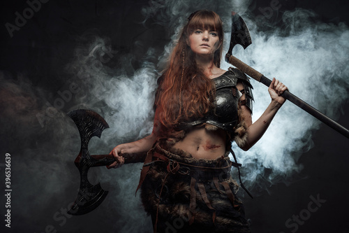 Portrait of beautiful and at the same time dangerous scandinavian woman fighter wielding two axes in dark smokey background.