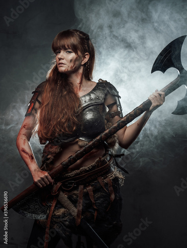 Portrait of woman viking wielding two handed axe and dressed in dark light armour in dark foggy background. © Fxquadro