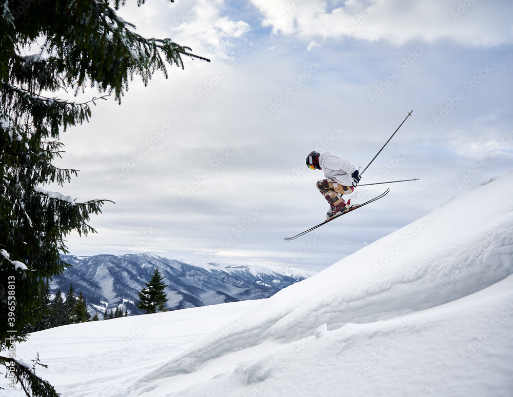 Side view of male skier making jump while sliding down snow-covered slope on skis under beautiful cloudy sky. Man freerider in helmet skiing on fresh powder snow in mountains.