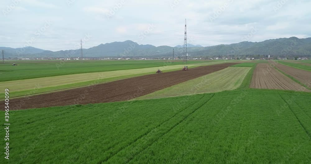 Aerial drone view of agriculture grassland field in summer. Tractors working on grassland meadow field. Beautiful landscape with small town and mountains in the distance. Backward moving Aerial drone view of agriculture grassland field in summer. Tractors working on grassland meadow field. Beautiful landscape with small town and mountains in the distance. Backward moving