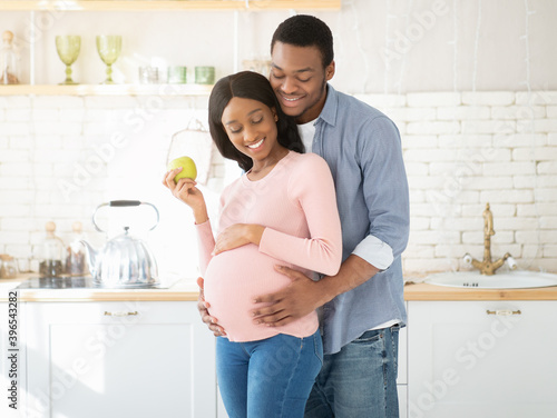 Portrait of loving black guy embracing his lovely expectant wife with apple in kitchen