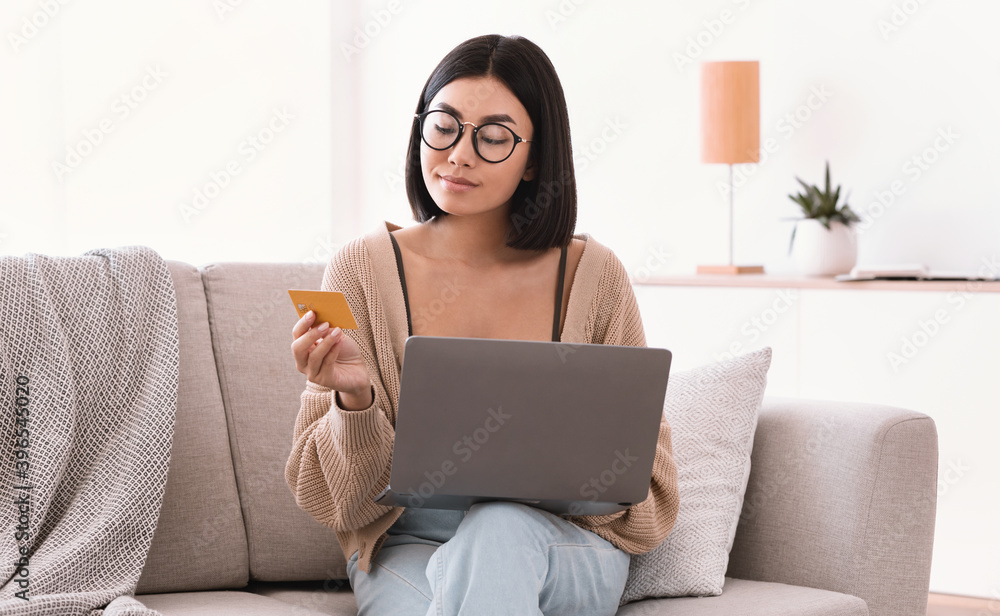Asian woman in glasses making purchases using laptop holding card Asian woman in glasses making purchases using laptop holding card