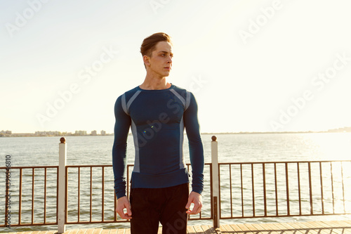 Fototapeta Naklejka Na Ścianę i Meble -  Healthy young male dressed in sportswear poses on pier. Sportsman walking by the sea at sunset. Photo of handsome athletic young man walking towards camera