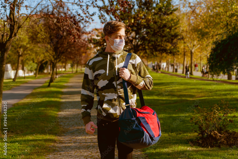 Obraz premium Young man going to the gym with his bag on his shoulder. wearing a protective mask is hiking through the woods. Young male athlete wearing a surgical mask is walking on the jogging track.