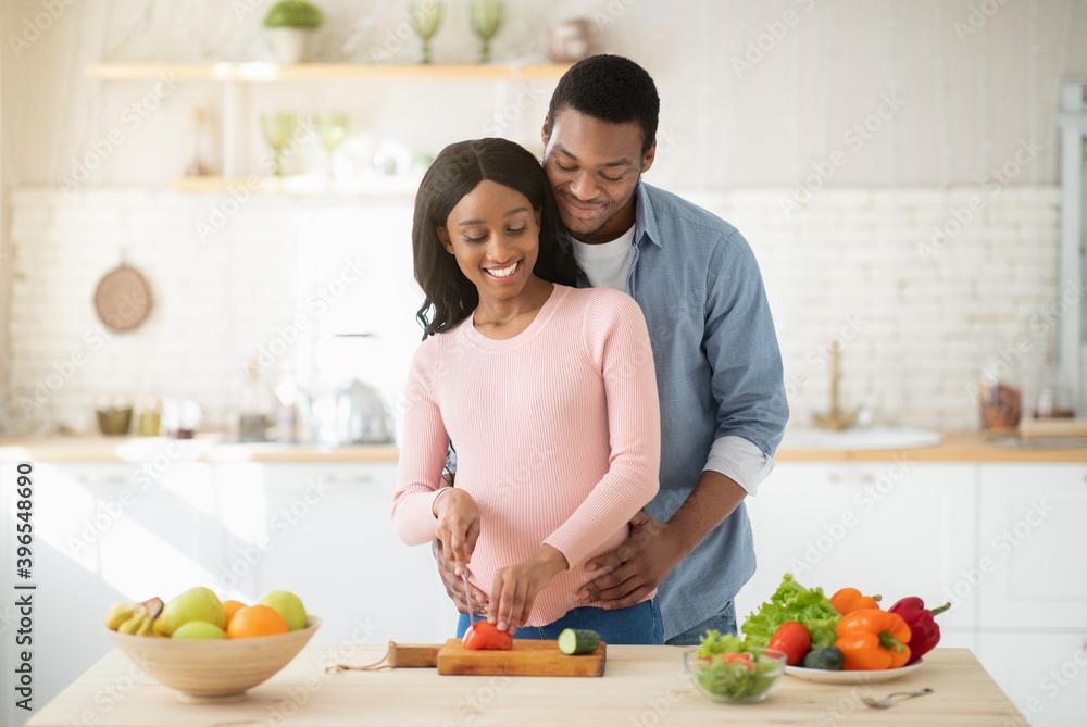 Happy young pregnant couple cooking fresh vegetable salad in kitchen, cutting tomato
