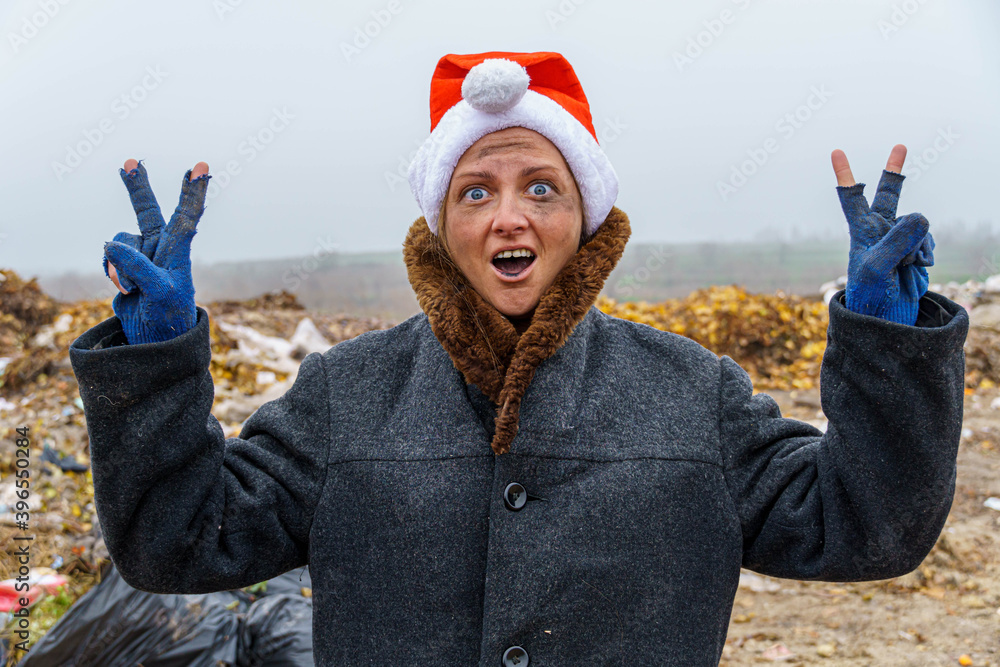 Obraz premium A dirty, poor woman in a New Year's hat, unsure of herself, shows two thumbs up against the background of a trash heap.