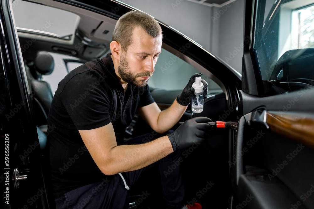 Young handsome car detailing worker making professional cleaning and remowing of the dust of car interior, using special cleansing disinfecting foam and soft brush. Car interior cleaning Young handsome car detailing worker making professional cleaning and remowing of the dust of car interior, using special cleansing disinfecting foam and soft brush. Car interior cleaning