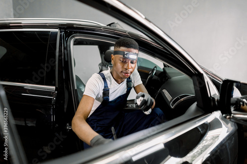 Front view of young black male worker in uniform, providing professional detailing cleaning of the interior and door of modern car, using special disinfecting foam and soft brush © sofiko14