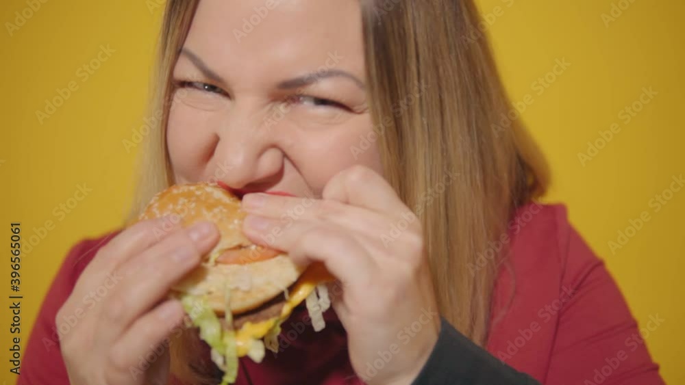 A plump woman greedily takes a bite of a burger. A plump woman greedily takes a bite of a burger.