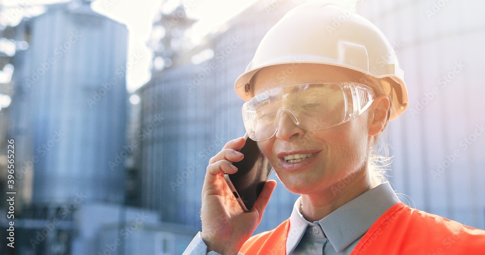 Close up portrait of joyful female engineer in goggles and helmet standing outdoors and chatting on cellphone. Woman builder having conversation on smartphone. Communication at work. Business concept Close up portrait of joyful female engineer in goggles and helmet standing outdoors and chatting on cellphone. Woman builder having conversation on smartphone. Communication at work. Business concept