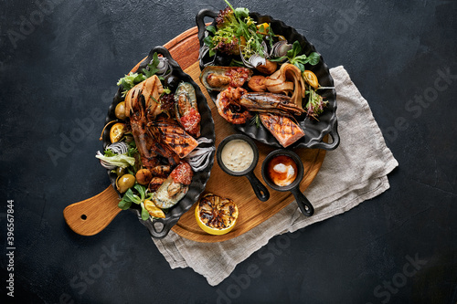 Mixed Roasted Seafood Platter Set. Contain Grilled Big Shrimps, Calamari Squids and Barracuda Fish Garlic Pepper with Spicy Chili Sauce and Potatoes, on wooden board and Black Background.