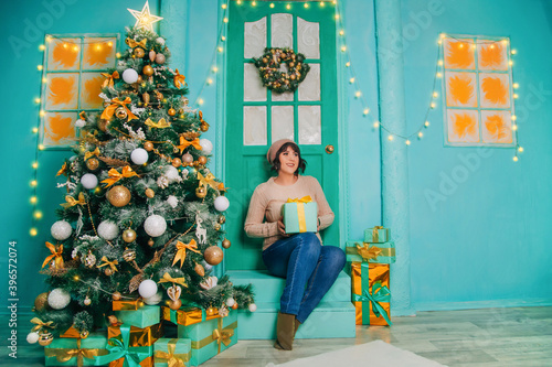 A girl in a white sweater sits on the porch at the Christmas door against the background of a Christmas tree with a gift. New Year's holidays. Snowing © Вероника Преображенс