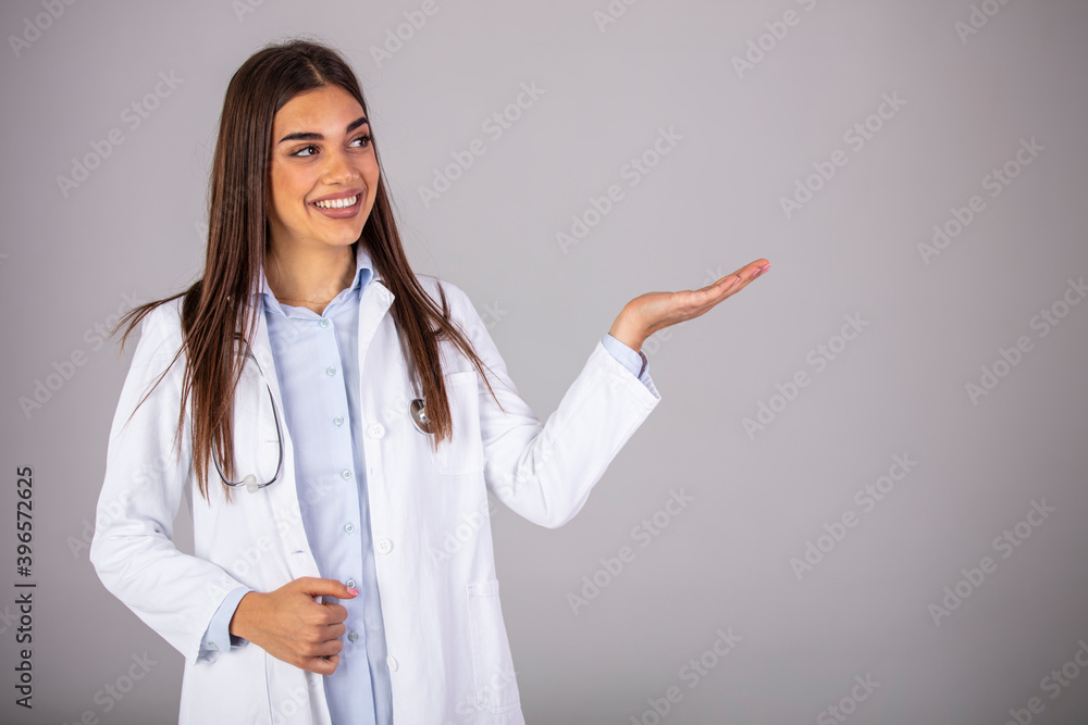 Smiling woman doctor showing aside to copyspace blank place. Confident female medical worker portrait pointing with finger to left side at empty space in studio isolated on gray background Smiling woman doctor showing aside to copyspace blank place. Confident female medical worker portrait pointing with finger to left side at empty space in studio isolated on gray background