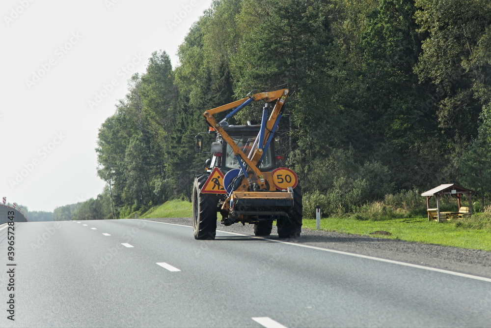 Wheeled tractor lawnmover with a external detachable mounted mower equipment movng on roadside of a suburban highway on forest background at summer day, road maintenance service mechanization machine Wheeled tractor lawnmover with a external detachable mounted mower equipment movng on roadside of a suburban highway on forest background at summer day, road maintenance service mechanization machine
