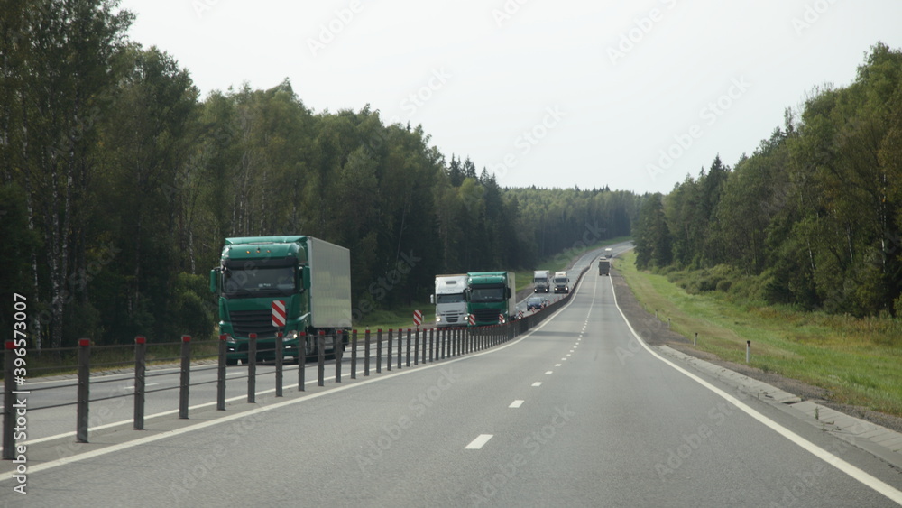 Semi trucks lorry convoy move on suburban asphalted highway road on fence and green forest on roadsides background front view at summer day,  on green forest background