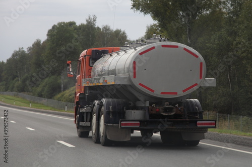Milk truck with a gray barrel drives on a suburban highway on a summer day, back side view on green forest and gray sky background, liquid food goods delivery