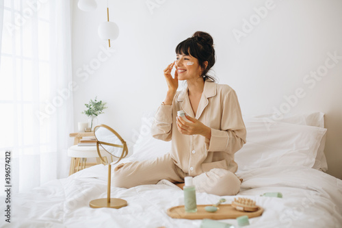 Smiling woman applying face cream sitting on bed