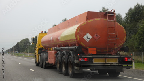 Modern orange semi truck fuel tanker with empty dangerous class sign on barrel move on suburban highway road at summer day in perspective, rear side view, car fuel ADR cargo transportation logistics