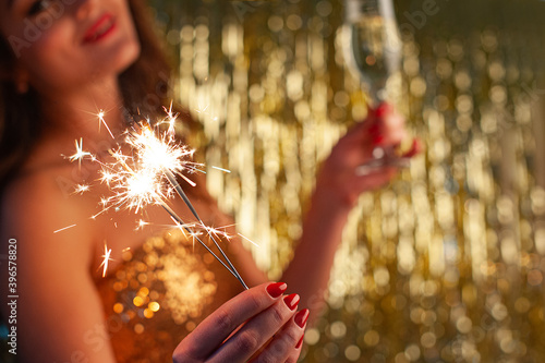 Obraz na płótnie Young woman drinks champagne and holding a sparkler on a bright gold background, selective focus w salonie