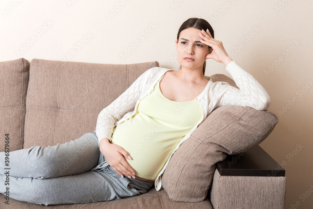 Young pregnant woman with headache sitting on sofa. Pregnancy expectation concept Young pregnant woman with headache sitting on sofa. Pregnancy expectation concept