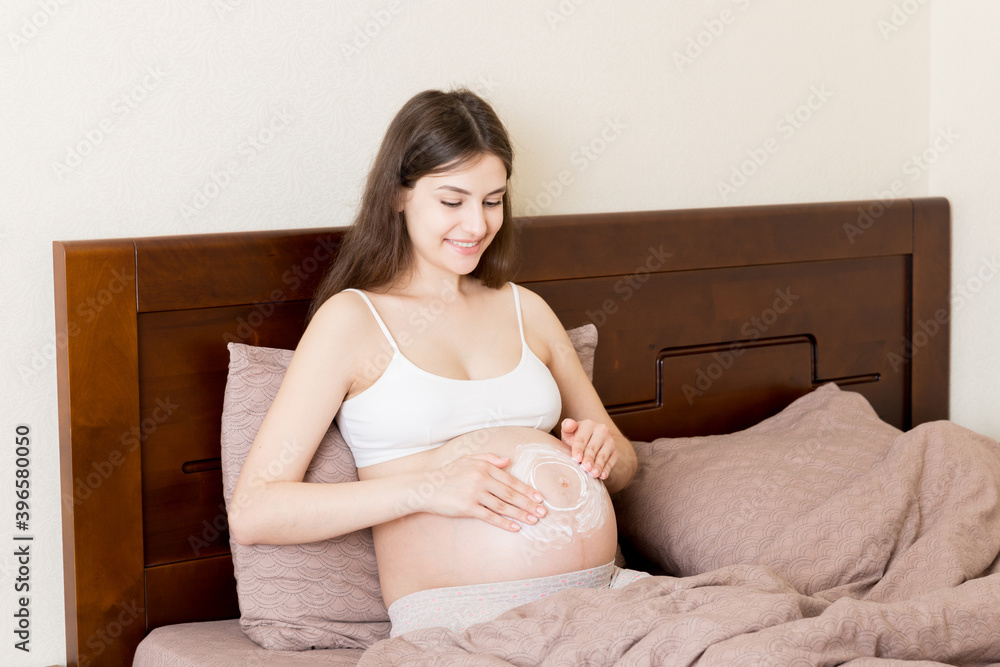 a pregnant girl sits at home on the bed and smears an anti-stretch mark cream on her stomach. Pregnancy, motherhood, preparation and expectation concept a pregnant girl sits at home on the bed and smears an anti-stretch mark cream on her stomach. Pregnancy, motherhood, preparation and expectation concept