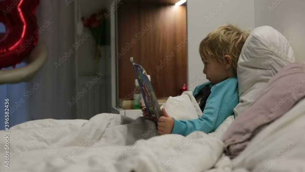 Sick toddler child, lying in bed with fever, reading a book