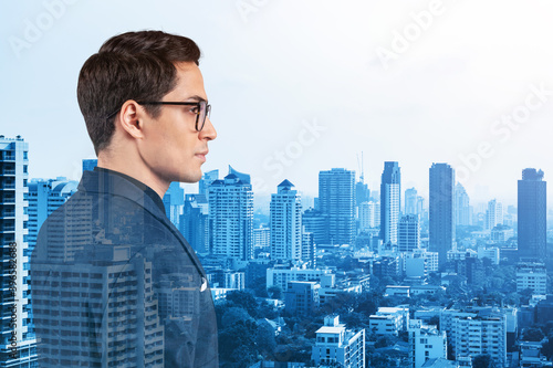 Young handsome businessman in suit and glasses dreaming about new career opportunities after MBA graduation. Bangkok on background. Double exposure.