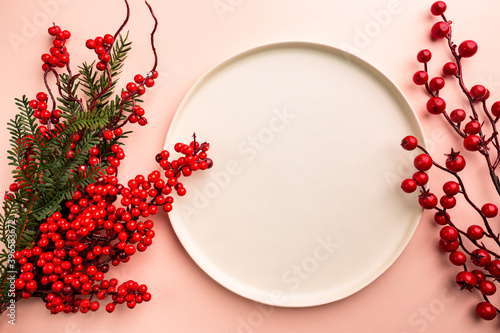 Christmas holidays composition with christmas decorations, red berries branches and white plate on pink background with copy space, top view